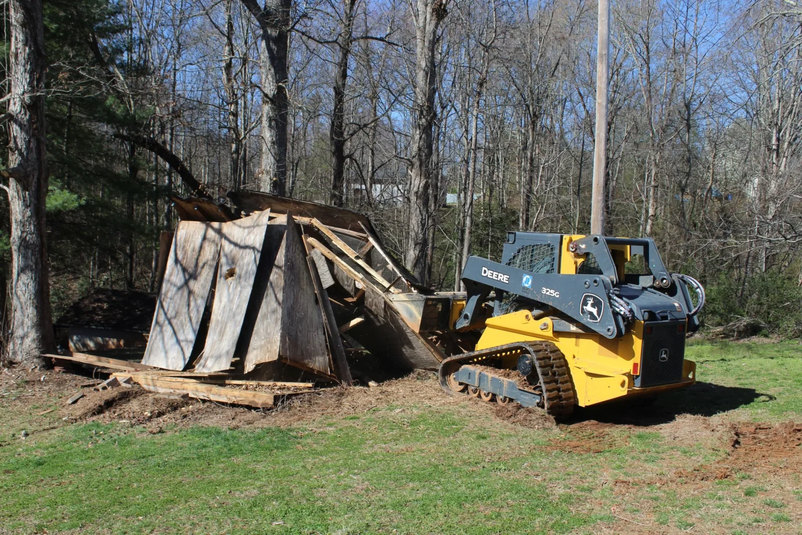 Compact track loader pushing an old plywood storage shed over at the edge of a wooded lot, the shed collapsing sideways as its roof panels fold